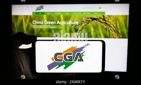 Person, die ein Mobiltelefon mit dem Logo des chinesischen Unternehmens China Green Agriculture Inc. (CGA) vor der Webseite hält. Schwerpunkt auf dem Display des Telefons. Stockfoto