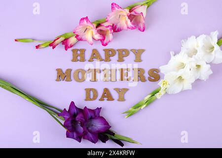 Grußkarte zum Muttertag. Weiße, lila und rosa Gladiolen Blumen auf einem lila Hintergrund mit einer Inschrift der Holzbuchstaben Muttertag. Stockfoto