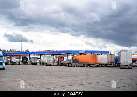 Industrietransporter verschiedene große Lkw-Sattelzugmaschinen mit beladenen Aufliegern stehen in Reihe auf der Tankstelle des Lkws und füllen Tanks bis Stockfoto