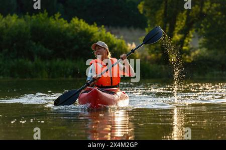 Lächelnde Frau, die während der goldenen Stunde auf einem ruhigen Fluss mit dem Kajak paddelt. Bild: Kajak Water Sports Concept Stockfoto