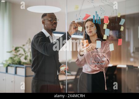 Junge Geschäftskollegen, die in einem modernen Büro mit Haftnotizen an einer Glaswand ein Brainstorming machen Stockfoto