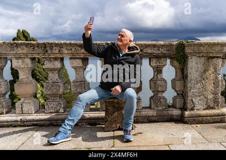 Glücklicher Reisender mittleren Alters im Urlaub, der ein Selfie auf einer Außenterrasse mit Blick auf den Lago Maggiore macht - Reise- und Urlaubskonzept Stockfoto