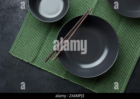 Leere japanische Gerichte. Eine schwarze Keramikschale für chinesische Nudeln oder Thai-Suppe liegt auf einem Bamkuk-Teppich. Weißer Topf für Sojasauce und rote chinesische Sticks Stockfoto
