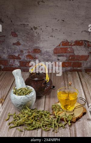 Zitronenkraut-Aufguss in einem Glasbecher mit einem Marmormörtel und Stößel und einer Keramik-Teekanne mit getrockneten Zitronenkraut-Blättern auf einem Holztisch mit einem Stockfoto