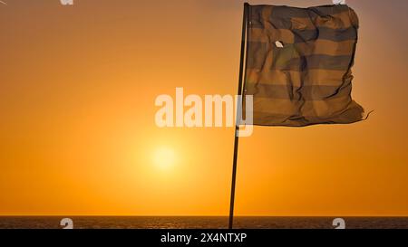 Eine griechische Flagge flattert im Wind vor einem leuchtend orangen Sonnenuntergang über dem Meer, Dämmerung, Sonnenuntergang, Rhodos, Dodekanesen, Griechische Inseln, Griechenland Stockfoto