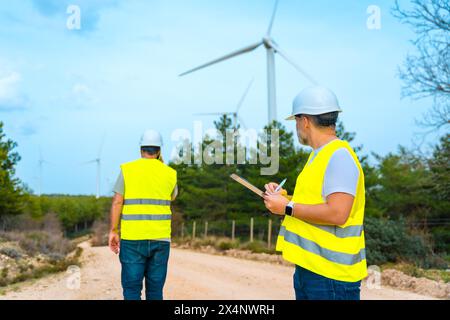 Techniker, die Windturbinen in einem grünen Energiepark inspizieren, sprechen mit dem Mobiltelefon und machen sich Notizen Stockfoto