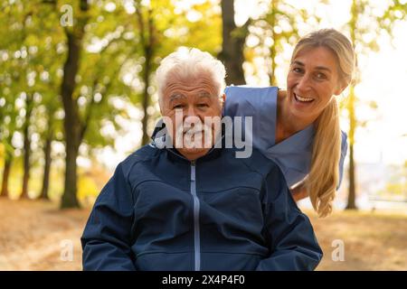 Ein älterer Mann im Rollstuhl, der einen freudigen Moment mit einer Krankenschwester in einem sonnendurchfluteten Park teilt, beide lächeln warm Stockfoto