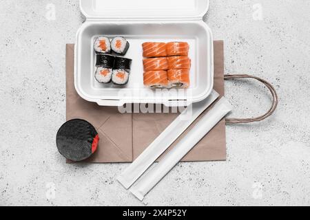 Plastikbehälter mit leckeren Sushi-Rollen, Essstäbchen und Papiertüte auf weißem Grunge-Hintergrund. Lieferkonzept Stockfoto
