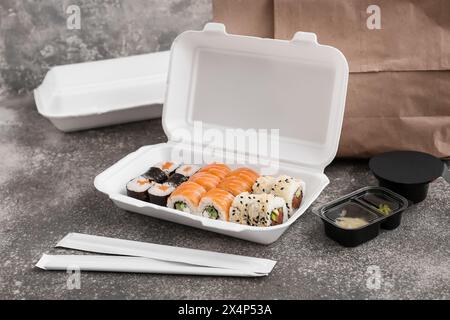 Plastikbehälter mit leckeren Sushi-Rollen, Essstäbchen und Papiertüte auf grauem Grunge-Hintergrund. Lieferkonzept Stockfoto