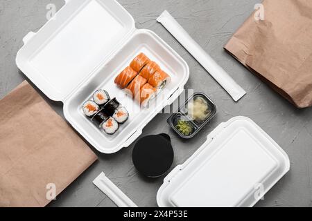 Plastikbehälter mit leckeren Sushi-Rollen, Essstäbchen und Papiertüten auf grauem Grunge-Hintergrund. Lieferkonzept Stockfoto