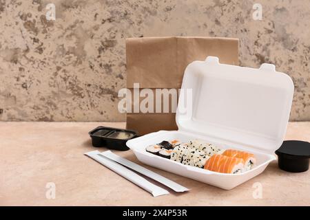 Plastikbehälter mit leckeren Sushi-Rollen, Essstäbchen und Papiertüte auf beigem Grunge-Hintergrund. Lieferkonzept Stockfoto
