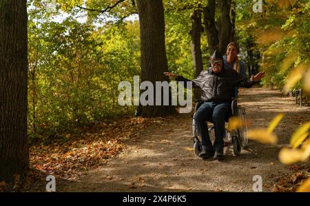 Großvater im Rollstuhl, gekleidet als Flieger, streckt freudig seine Arme, umgeben von bunten Herbstbäumen Stockfoto