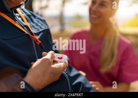 Ein Senior Mann hält einen SOS-Alarm-Knopf an einem Schlüsselband, während ihn eine Pflegekraft anlächelt, verschwommener Herbsthintergrund Stockfoto