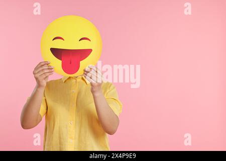 Frau, die das Gesicht mit Emoticon bedeckt und die Zunge auf rosa Hintergrund hinausragt. Leerzeichen für Text Stockfoto