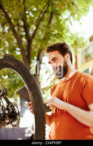 Enthusiastischer gesunder männlicher Radfahrer, der Reifen als jährliche Fahrradgummi-Wartungsroutine mit digitalem Tablet überprüft. Kaukausischer Mann, der intelligente Geräte benutzt, um nach Lösungen für beschädigte Fahrradreifen zu suchen. Stockfoto