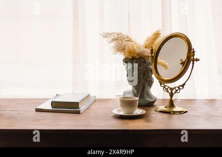 Eleganter Vintage-Spiegel und Büstenvase mit Pampas-Gras neben einer Kaffeetasse auf einem Holztisch Stockfoto