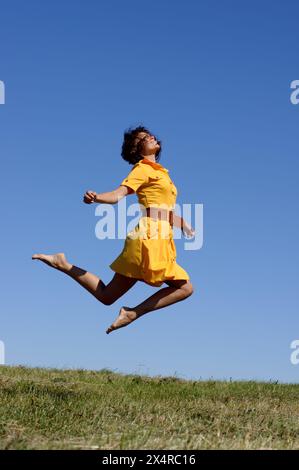 Glückliche junge Frau in gelbem Kleid, die auf dem grünen Feld springt Stockfoto