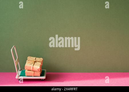 Trolley-Wagen mit Geschenkboxen auf rosa und khakigrünem Hintergrund. Paketzustellungskonzept Stockfoto