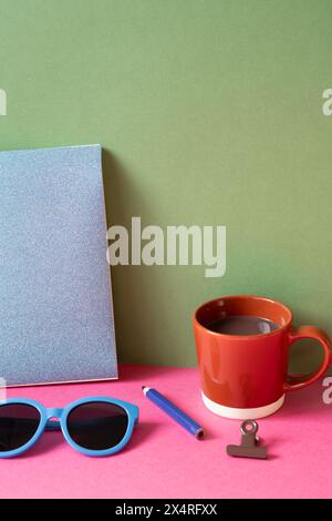 Desktop-Arbeitsbereich. Notizbuch, Brille, Farbstift, Clip, Tasse heiße Schokolade auf rosa Schreibtisch. Khakifarbener grüner Wandhintergrund Stockfoto