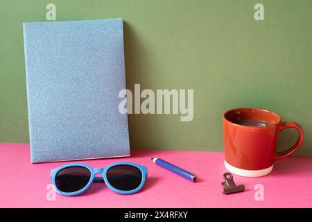 Desktop-Arbeitsbereich. Notizbuch, Brille, Farbstift, Clip, Tasse heiße Schokolade auf rosa Schreibtisch. Khakifarbener grüner Wandhintergrund Stockfoto