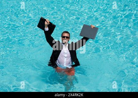 Ein lustiger, überraschter Geschäftsmann im Anzug mit einem Laptop im Swimmingpool. Verrückter, aufgeregter Geschäftsmann im Sommerurlaub. Aufgeregter Geschäftsmann in Neoprenanzug aufgeregt Stockfoto