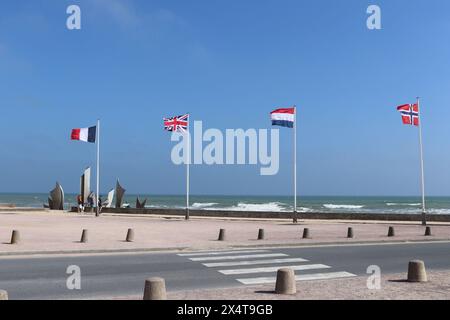 Blick auf die Flaggen und das Denkmal am Strand von Omaha. Wellen, die vom Ozean hereinrollen. Blauer Himmel und ein paar sichtbare Menschen. Stockfoto