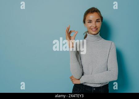 Eine bewusste Frau in einem grauen Pullover bildet ein perfektes „OK“-Zeichen, das ihr Selbstvertrauen eine elegante Haltung ergänzt. Stockfoto
