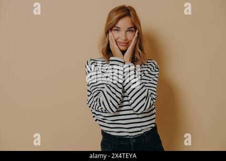 Hände rahmen ein strahlendes Lächeln, während eine Frau in Streifen eine verspielte Aura auf einer beigen Leinwand zeigt. Stockfoto