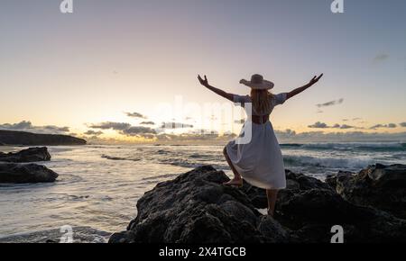 Rückansicht einer Frau in weißem Kleid und Strohhut mit erhobenen Armen, stehend auf Felsen bei Sonnenuntergang am Meer Stockfoto