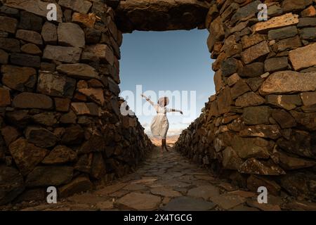 Eine Frau in weißem Kleid und Strohhut springt freudig in einen Steinbogen auf fuerteventura Stockfoto