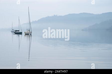 Atemberaubendes, friedliches Landschaftsbild des nebeligen Frühlingsvormittags über Windermere im Lake District mit Booten, die auf dem See verankert sind, und entfernten nebeligen Gipfeln Stockfoto