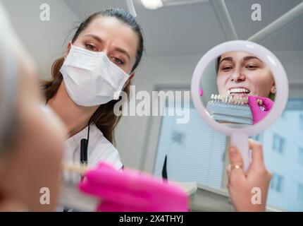 Junge Frau sitzt im Stomatologie-Klinikstuhl und lächelt im Spiegel, zeigt ihre Zähne und wählt Zahnfarben mit Farbtabellen. Gesundheit, Mund Stockfoto