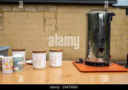 Eine verchromte elektrische Warmwasserurne für englischen Tee, Töpfe mit Tee und Zucker und Tassen auf einem befleckten Tisch. Stockfoto