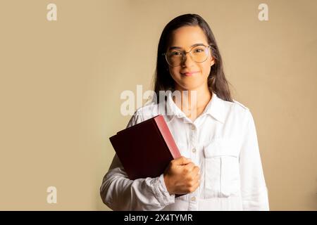 studio-Foto einer jungen Latina, die ein Buch hält, lächelt und geradeaus blickt, auf einem neutralen Hintergrund Stockfoto