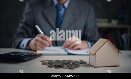 Ein Agent in Anzug macht Notizen nach Hausmodell und Münzstapeln, schlägt Immobilieninvestitionen vor oder schreibt einen Vertrag und versicherst den Kauf und Verkauf von Eigenheimen. busin Stockfoto