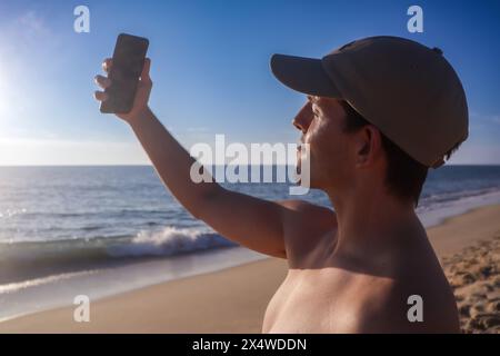 Junger Mann mit einer Mütze, der lächelt, während er am Strand ein Selfie mit Sonnenuntergang im Hintergrund macht Stockfoto