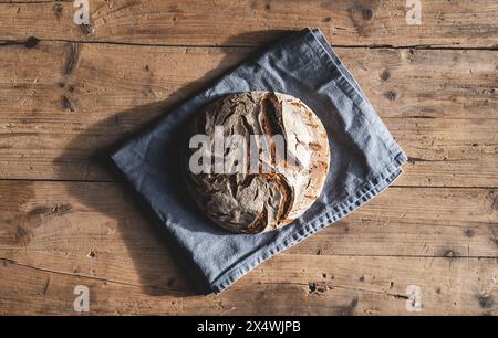 Frisch gebackener runder Brotlaib auf einem blauen Tuch, auf einem rustikalen Holztisch Stockfoto
