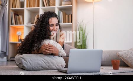 Wochenendfilm. Gemütliche Freizeit. Amüsantes, entspanntes lächelndes Mädchen, das Spaß an einer lustigen Videoshow online auf einem Laptop auf dem Boden in hellem Wohnraum mit freiem Platz genießt. Stockfoto