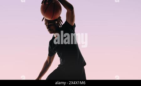 Ein junger Athlet mit Dreadlocks dribbelt einen Basketball in einem Studio. Seine Form ist auf den Punkt gekommen, während er beleidigt, zeigt Geschick und Leichtathletik. Stockfoto