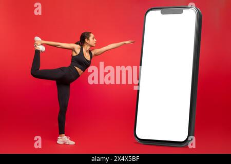 Flexible junge Frau in schwarzer Sportswear, die neben dem großen Handy trainiert Stockfoto