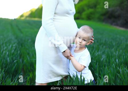 Eine Schwangerschaft, die mit einem Jungen auf einem Feld steht. Ich erwarte mein drittes Kind. Die Frau trägt ein weißes Kleid und hat ihre Hände auf der Schulter o Stockfoto