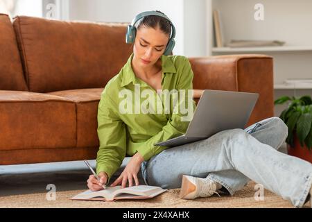 Junge Frau, die Kopfhörer trägt, während sie Notizen schreibt und Laptop benutzt Stockfoto