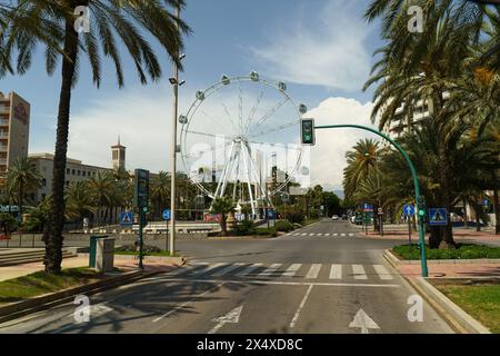 Almeria, Spanien - 25. Mai 2023: Eine von hohen Palmen gesäumte Straße führt zu einem riesigen Riesenrad in der Ferne, unter einem klaren blauen Himmel. Stockfoto