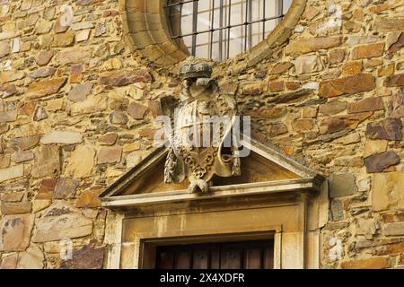 Stilisiertes Steinwappen mit komplizierten Details ziert die Außenwand über einer Holztür und fügt sich in die rustikale Textur der Steinziegel ein. Stockfoto