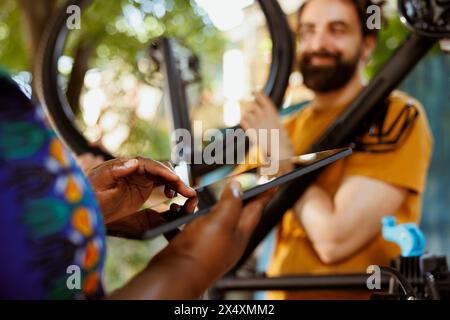 Nahaufnahme einer schwarzen Frau, die ein Handy-Tablet greift, während ein enthusiastischer junger Mann im Hintergrund das Fahrrad im Freien repariert. afroamerikanerin, die ein digitales Gerät hält, um Reparaturen für kaputte Radreifen zu untersuchen. Stockfoto