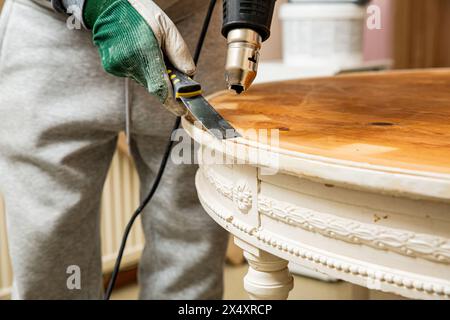 Mann mittleren Alters, der alten Holztisch mit einer Heißluftpistole hochfährt, um alte Schmerzen zu beseitigen. Möbelsanierung, Heimwerkerreparatur, Reparatur durch sich selbst und Improvisation Stockfoto