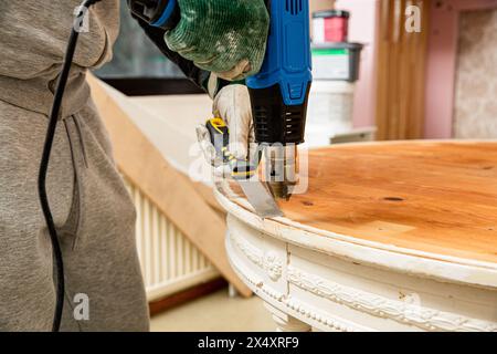 Mann mittleren Alters, der alten Holztisch mit einer Heißluftpistole hochfährt, um alte Schmerzen zu beseitigen. Möbelsanierung, Heimwerkerreparatur, Reparatur durch sich selbst und Improvisation Stockfoto