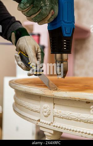Mann mittleren Alters, der alten Holztisch mit einer Heißluftpistole hochfährt, um alte Schmerzen zu beseitigen. Möbelsanierung, Heimwerkerreparatur, Reparatur durch sich selbst und Improvisation Stockfoto