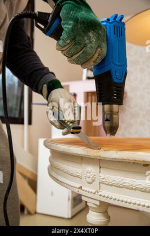 Mann mittleren Alters, der alten Holztisch mit einer Heißluftpistole hochfährt, um alte Schmerzen zu beseitigen. Möbelsanierung, Heimwerkerreparatur, Reparatur durch sich selbst und Improvisation Stockfoto