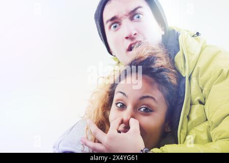 Humorvolle und offene Szene mit einem jungen Mann in einer Mütze und gelber Jacke, die spielerisch den Mund einer Frau bedeckt, beide mit verspielten Ausdrücken Stockfoto